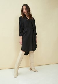Zwarte shirtjurk met een ceintuur, lange mouwen en een hoge-lage zoom, gecombineerd met crème kniehoog hakken. Effen textuur en minimalistisch ontwerp.