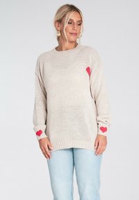 Beżowy sweter dziany z czerwonymi akcentami w kształcie serca na rękawach, prążkowany dekolt i mankiety. W komplecie z jasnoniebieskimi dżinsami.