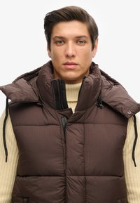 Braune Weste mit hoher Kragen, schrägen Nähten und Reißverschlusstaschen. Kombiniert mit einem strukturierten cremigen Pullover darunter.