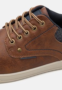 Chaussure en cuir marron avec semelle en caoutchouc beige, œillets dorés et lacets tressés. Présente un accent en tissu gris et un détail de couture bleu.