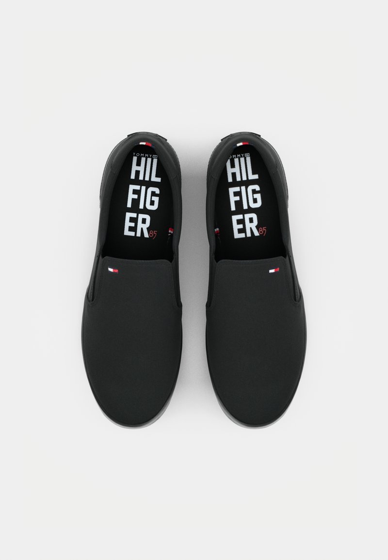 Baskets noires à enfiler avec une texture lisse, dotées d'une semelle intérieure de marque et d'un petit accent de logo en rouge et blanc. Design minimaliste.