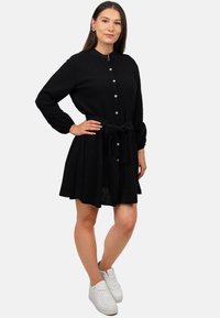 Abito nero a camicia realizzato in tessuto testurizzato, con maniche lunghe, una cintura a cravatta in vita e una gonna svasata. Abbinato a sneakers bianche.