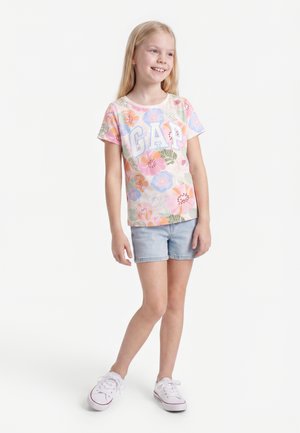 Fille souriante aux longs cheveux blonds portant un t-shirt GAP fleuri pastel, un short en jean clair et des baskets blanches, debout sur un fond blanc.
