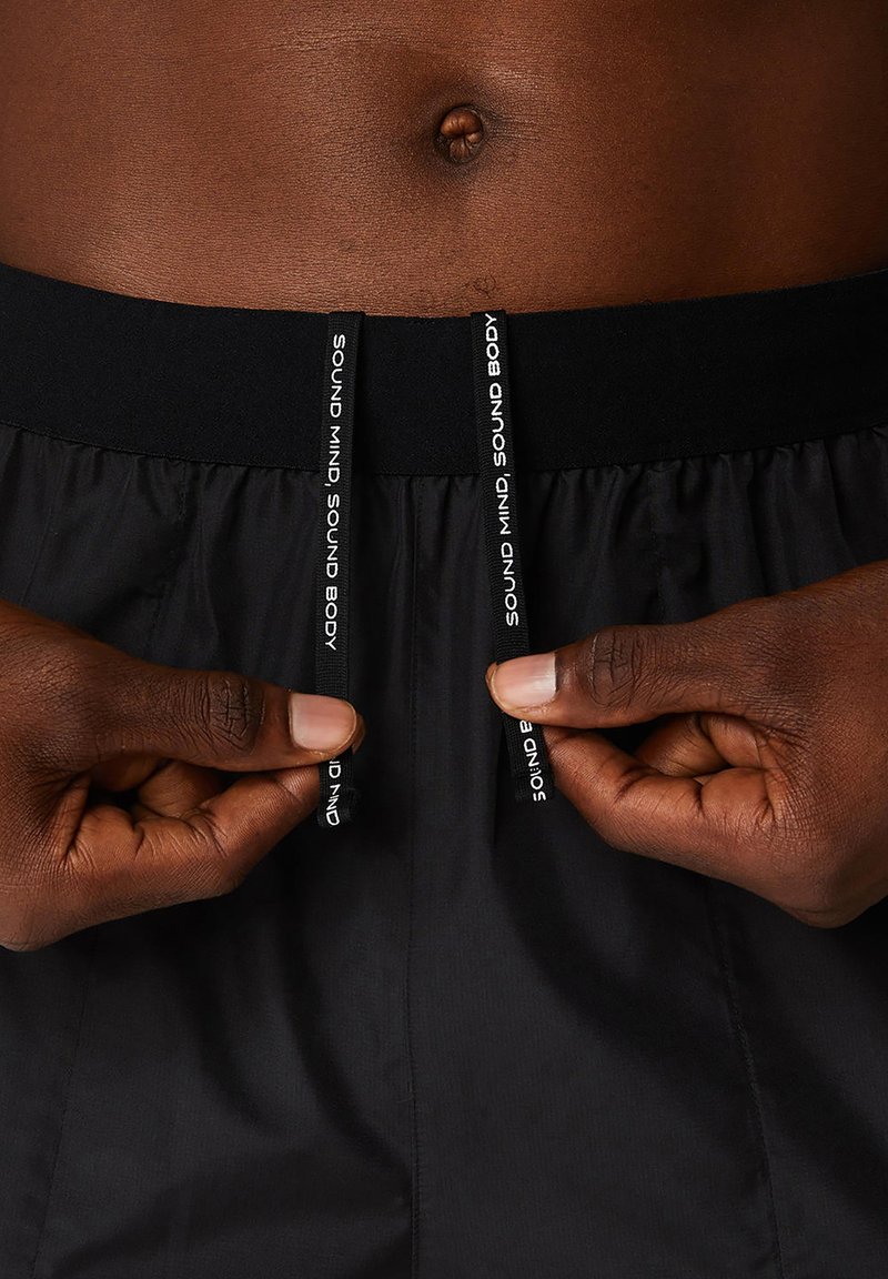 Shorts de sport noirs avec cordons de serrage. La taille est élastique ; les cordons de serrage portent le texte "SOUND MIND SOUND BODY" en blanc.