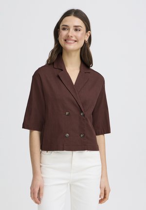 Ung kvinde smilende, iført en brun kortærmet dobbeltradet jakke og hvide bukser, stående mod en ensfarvet lys baggrund.