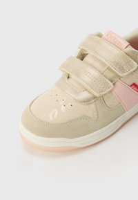 Adidași bej cu accente din suede și lucioase, având două benzi Velcro și detalii roz. Vârf rotund cu orificii de ventilație. Talpă din cauciuc.