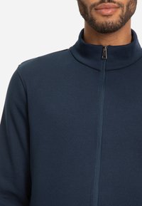 Chaqueta azul marino con cremallera y cuello alto, hecha de una tela suave, que presenta una cremallera central y costuras decorativas a lo largo de las costuras.