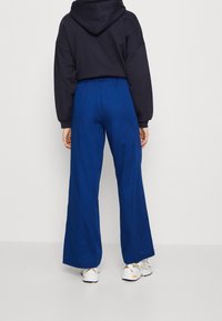 Námořnická mikina s kapucí na stažení a volným střihem, doplněná širokými, jasně modrými kalhotami s širokými nohavicemi. Na dně jsou částečně viditelné bílé sportovní boty.