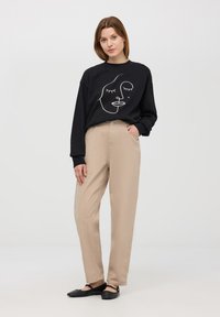 Felpa nera con design di un volto in linea bianca, abbinata a pantaloni beige a gamba dritta. Scarpe nere flat completano l'outfit. Tessuto dalla texture liscia.