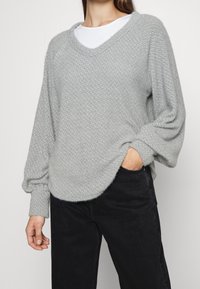 Pull en maille texturée gris clair avec un col en V et des épaules tombantes, offrant une coupe décontractée et des manches longues. Associé à un pantalon noir.