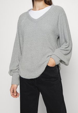 Maglione a maglia testurizzata di un grigio chiaro, con scollatura a V e spalle scese, caratterizzato da una vestibilità rilassata e maniche lunghe. Abbinato a pantaloni neri.