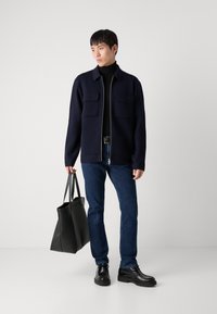 Mornarska zip-up jakna z dvema žepoma na prsih, črna turtleneck majica, modri kavbojki, črne usnjene čevlje in črna torba.