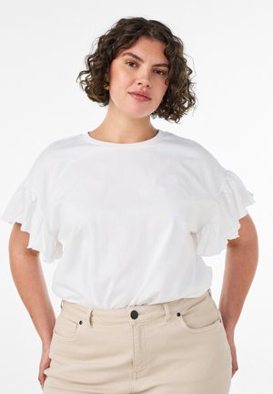 Kvinde med krøllet hår iført hvid rynket kortærmet top, stoppet ned i beige bukser med høj talje, poserer foran en hvid baggrund.