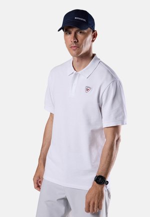 Mann trägt ein weißes Poloshirt mit kleinem Logo, hellgraue Hose, eine schwarze Armbanduhr und eine marineblaue Kappe mit weißem Schriftzug.