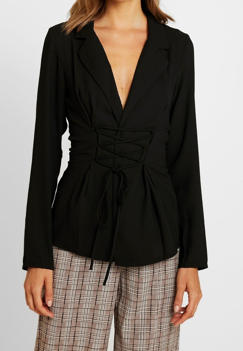 Femme portant un blazer noir à manches longues avec un détail de laçage à l'avant et un pantalon large à carreaux beige, debout devant un fond uni.