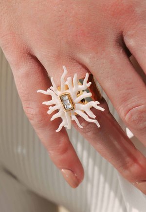 Bague en forme de corail blanc avec des accents dorés et une pierre rectangulaire transparente, portée sur une main à la peau claire et avec un vernis à ongles neutre.