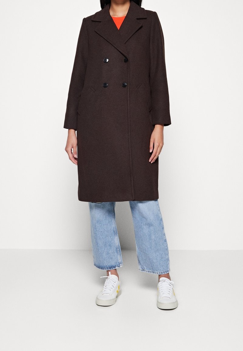 Hnědý kabát s dvojitým zapínáním z vlněné směsi s černými knoflíky, velkými klopami a bočními kapsami, v kombinaci se světle modrými džíny a bílými teniskami.