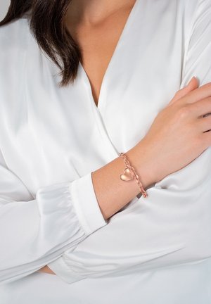 Bracciale in oro rosa con un ciondolo a forma di cuore, caratterizzato da un design a catena intrecciata. Indossato su un polso, abbinato a una maglietta bianca a maniche lunghe.