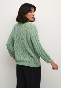 Light green knitted sweater with a loose fit, featuring a wave pattern, long sleeves, and ribbed cuffs and hem. Back view shows open weave detail.