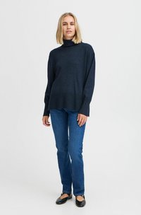 Dunkelblauer Rollkragenpullover mit entspannter Passform, geripptem Saum und langen Ärmeln, kombiniert mit blauen Jeans und schwarzen Ballerinas. Glatte Textur.
