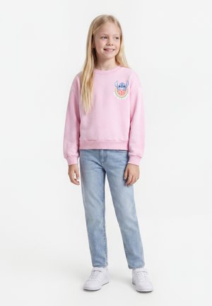 Νεαρή κοπέλα με μακριά ξανθά μαλλιά, που φοράει ένα ροζ φούτερ με ένα μπλε καρτούν χαρακτήρα και καρπούζι, ανοιχτό μπλε τζιν και λευκά αθλητικά παπούτσια.