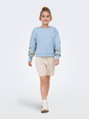Jeune fille marchant, portant un sweat bleu clair avec des motifs floraux sur les manches, un short beige, des chaussettes blanches et des baskets blanches.