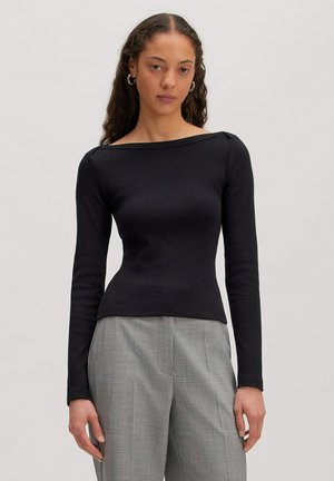 Frau mit langen lockigen Haaren, die ein schwarzes Langarmoberteil und graue, hoch taillierte Hosen trägt, stehend vor einem schlichten hellen Hintergrund.