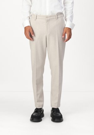 Pantalon beige ajusté avec une coupe droite, texture subtile, deux poches avant et fermeture à bouton, associé à des chaussures noires.