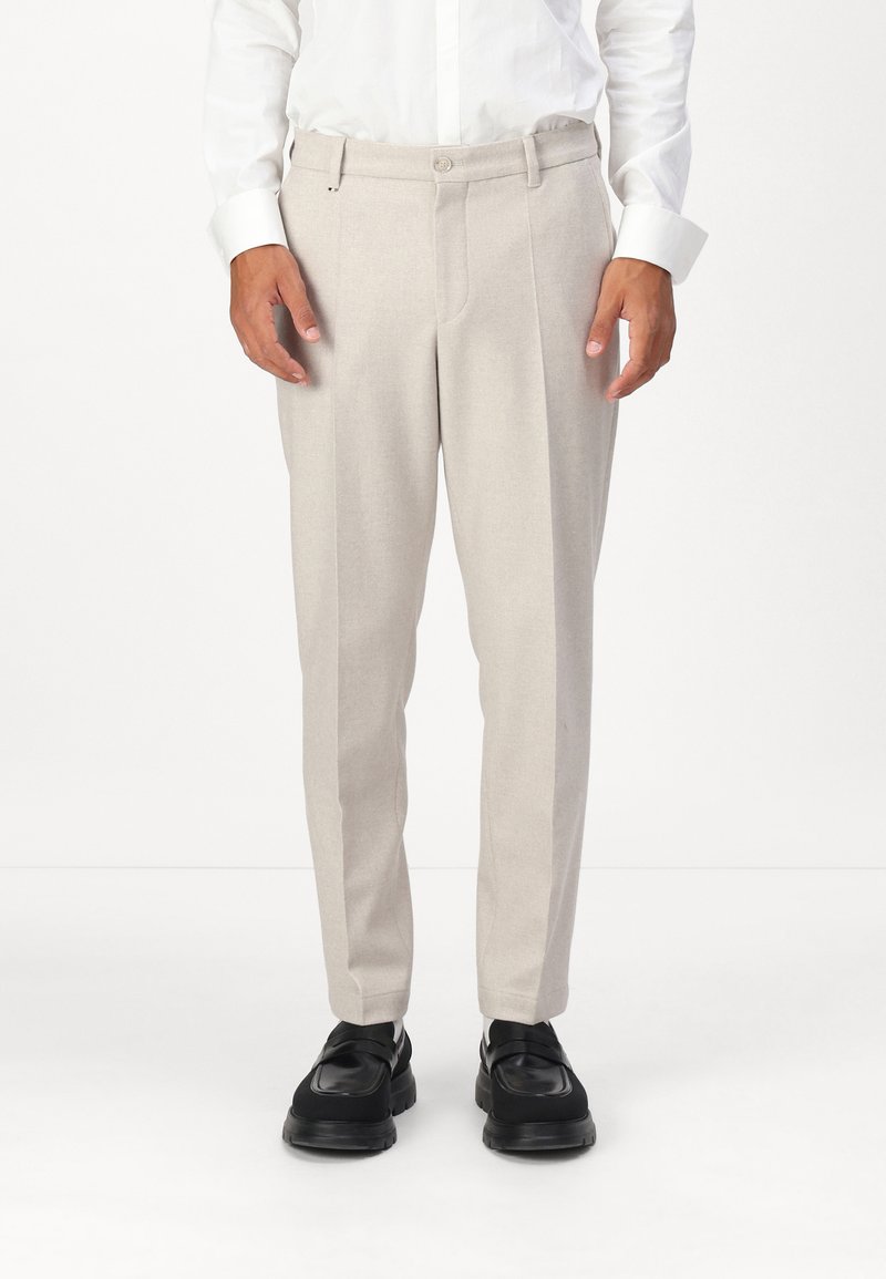 Beige maßgeschneiderte Hose mit geradem Schnitt, subtiler Textur, zwei Fronttaschen und Knopfverschluss, kombiniert mit schwarzen Schuhen.