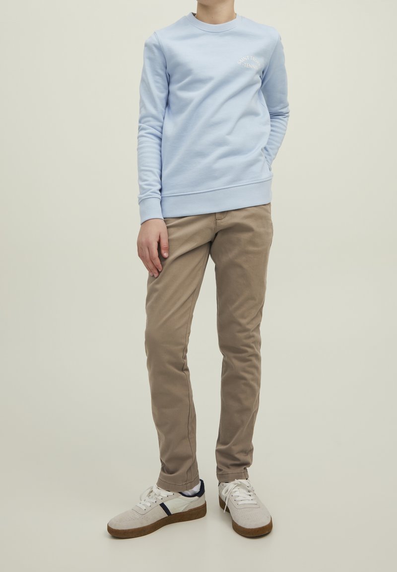 Personne portant un sweat-shirt bleu clair avec le texte "Saint Tropez Tennis", un pantalon beige et des baskets blanches à semelles marron, debout de manière détendue.