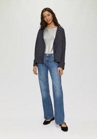 Dunkelblauer Cardigan mit Rippensaum, silbernem Metallic-Oberteil und hellblauen Schlagjeans. Das Modell trägt schwarze Ballerinas. Einfacher Hintergrund.