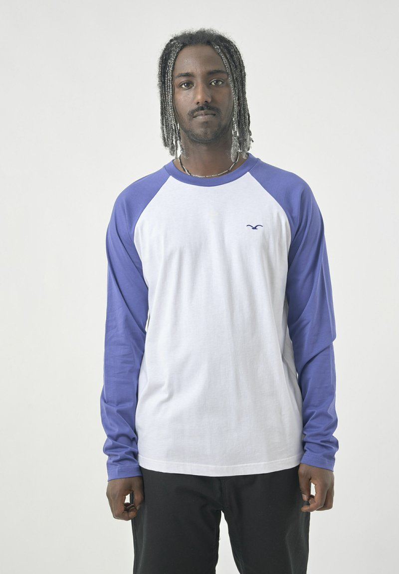 Langärmliges Raglan-T-Shirt mit weißem Körper und lila Ärmeln. Weiches Baumwollmaterial mit einem dezenten Logo auf der Brust.