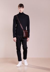 Borsa a tracolla in pelle marrone con una texture liscia e chiusura con cerniera, indossata da un modello con un dolcevita nero e pantaloni neri.