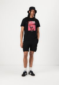 T-shirt en coton noir avec un grand imprimé graphique rose, associé à un short noir et des baskets noires avec des rayures blanches.