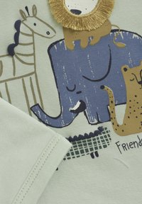 Ein hellgrüner Stoff zeigt Illustrationen eines blauen Elefanten, eines Löwen, einer Giraffe und eines braunen Hundes mit schwarzen Akzenten, beschriftet mit "Freunde".