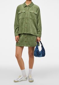 Camicia di velluto a coste verde con due tasche sul petto, abbinata a una gonna coordinata. Sneakers argento e una borsa a tracolla blu con motivo completano l'outfit.