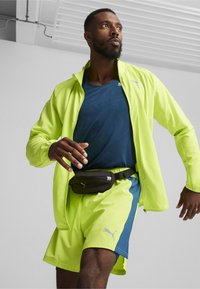 Tenue de sport comprenant une veste néon jaune vif et un short, assortis d'un t-shirt bleu foncé et d'une ceinture noire, conçue pour le mouvement.