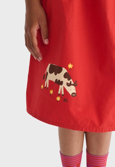 Main reposant à côté d'une jupe rouge avec un imprimé de vache noir et blanc et des fleurs jaunes au-dessus de chaussettes rayées roses et violettes sur les jambes.