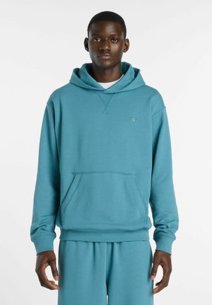 Mann trägt einen petrolblauen Kapuzenpullover und passende Hose und steht vor einem schlichten weißen Hintergrund.