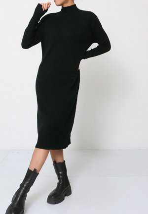 Femme posant en robe midi noire à col roulé et manches longues, avec des bottines à talons carrés en cuir noir, sur fond blanc.