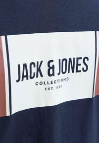 Sötétkék póló, nagy fehér téglalap alakú grafikával, rajta vastag fekete betűkkel "JACK & JONES" felirattal és barna díszítéssel. Texturált anyag.