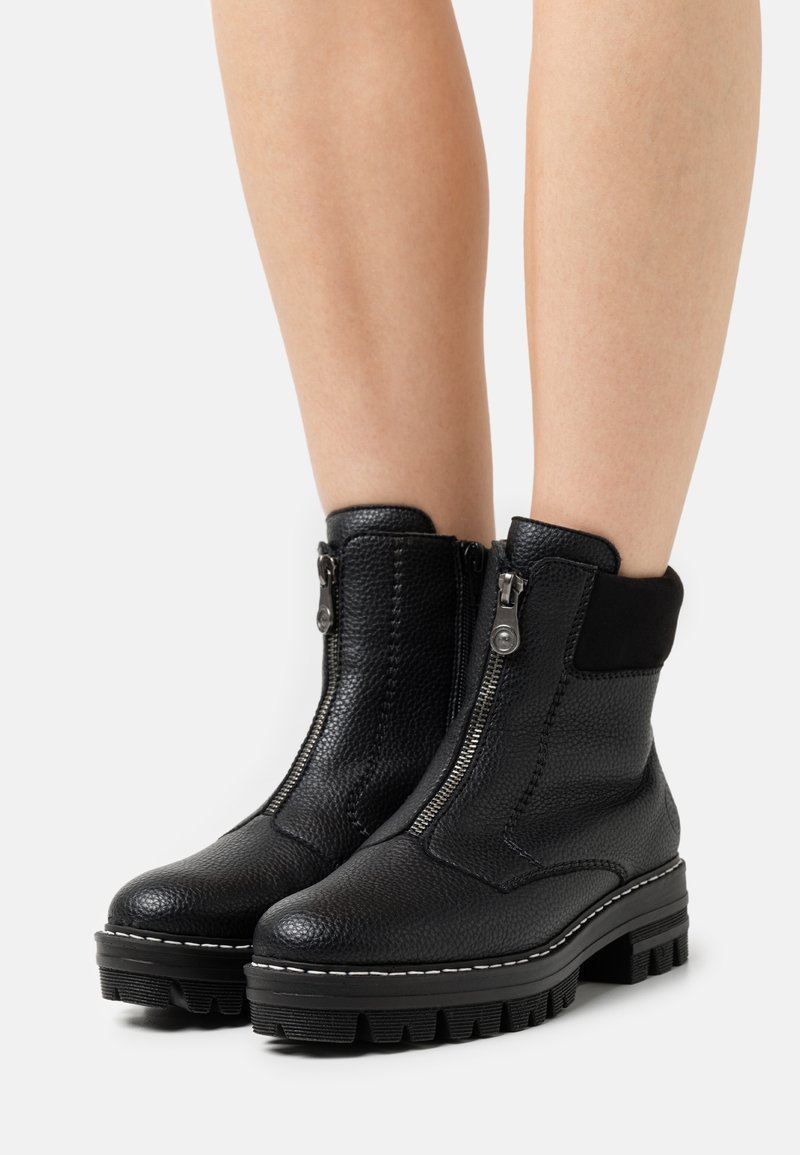 Bottes noires en cuir texturé à cheville avec fermetures éclair frontales, semelles épaisses à crampons, et collets rembourrés portées sur des jambes nues sur un fond blanc.