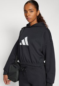 Juodas džemperis su baltu Adidas logotipu, pagamintas iš minkštos medžiagos. Su elastinga juosmens juosta ir šoninėmis kišenėmis. Derinamas su maža juoda kuprine.