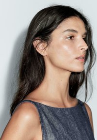 Giovane donna con lunghi capelli scuri che indossa un vestito di jeans senza maniche, rivolta a destra su uno sfondo grigio chiaro uniforme.