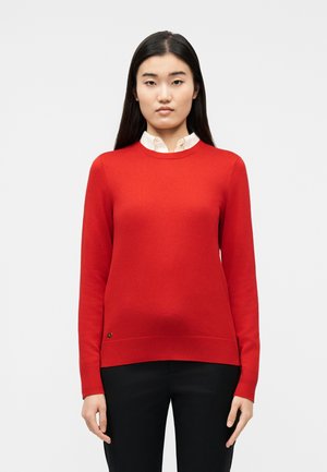 Femeie cu păr lung negru, purtând un pulover roșu cu gât rotund peste o cămașă albă cu guler și pantaloni negri, stând pe un fundal simplu.