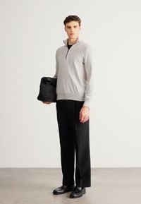 Halli tõmblukuga kampsun peene logoga, sobitatud mustade pükstega. Modell hoiab musta kotti ja kannab musti kingi. Lihtne, minimalistlik disain.