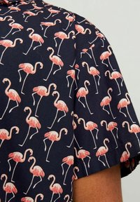 Camisa de manga corta con fondo azul marino, que presenta un patrón de flamencos rosas repetido. El material parece ligero y tiene una textura suave.