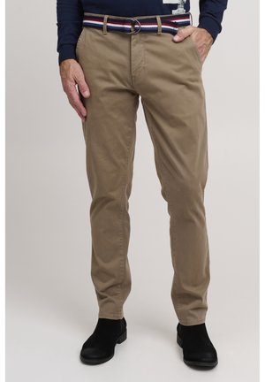 Man draagt beige strakke broek, zwarte enkellaarzen, gestreepte riem en een marineblauw shirt met lange mouwen, hand in de zak.