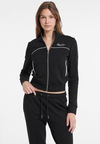Felpa cropped nera con zip, dettagli in bianco, maniche lunghe e vita aderente. Presenta un piccolo logo ricamato sul petto.