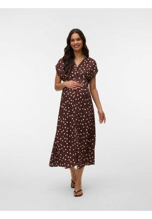 Femme enceinte souriante en robe midi marron à pois blancs, tenant son ventre, marchant vers l'avant avec des sandales noires ouvertes aux orteils.